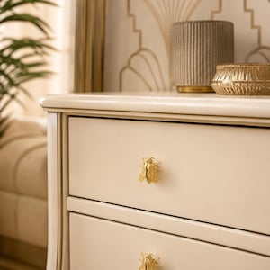 May include: A cream-colored wooden dresser featuring two drawers, each equipped with a detailed gold-toned bee-shaped metal knob. The dresser has a smooth, matte finish and a classic rectangular frame with subtle decorative molding along the edges.