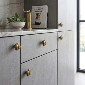 Pode incluir: Um armário de cozinha com portas cinzentas e ferragens de latão. Um moedor de café e um livro intitulado "Coffee Brewing" estão sobre a bancada. Uma pequena tigela branca com uma planta verde também está na bancada.