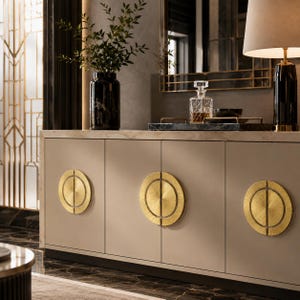 Puede incluir: Un aparador beige con tres puertas, cada una con un gran tirador circular dorado. El aparador tiene una superficie de mármol y un jarrón negro con vegetación encima. Un espejo y una lámpara son visibles en el fondo.