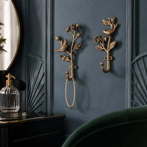 May include: Two wall-mounted hooks made of antique gold-toned metal, each featuring a sculpted branch design with leaves, flower buds, and a small bird perched on the stem. A delicate white beaded necklace hangs from the left hook.