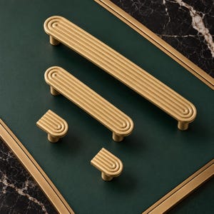 May include: A collection of four gold-toned metal cabinet hardware pieces, including two long rectangular pulls and two smaller semi-circular knobs. Each piece features a series of concentric, raised linear ridges along the top surface.