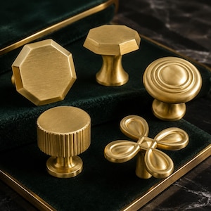 May include: A collection of five brass cabinet knobs with a brushed gold finish. The set includes two octagonal knobs, one circular knob with concentric rings, one cylindrical knob with vertical fluting, and one four-petaled floral shaped knob.