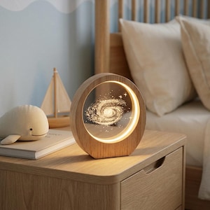 May include: A circular wooden lamp featuring a transparent center panel with an illuminated, laser-engraved spiral galaxy design. The light emits a warm glow from within the circular frame, highlighting the intricate star and galaxy details.
