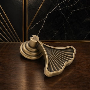 Può includere: Una maniglia per armadio color bronzo con un design a foglia di ginkgo. La maniglia presenta una base rotonda con dettagli a spirale e una maniglia a forma di ventaglio con scanalature lineari. L'hardware è posizionato su una superficie in legno scuro e uno sfondo in marmo nero.