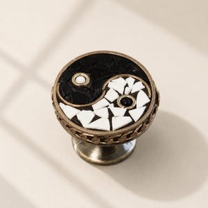 Peut inclure: Un bouton de meuble rond arborant un symbole yin et yang composé de carreaux de mosaïque noirs et blancs. La face circulaire est encadrée par une bordure en métal bronze à motif de maillons de chaîne, avec une perle blanche et une perle noire.