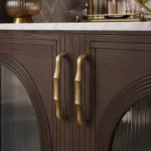 May include: A dark brown wooden cabinet featuring two vertical doors with arched, fluted glass panels. The doors are equipped with two long, curved, antique brass-toned metal handles. The cabinet is topped with a white marble surface.