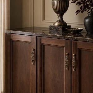May include: A dark brown wooden cabinet featuring three doors with vertical fluted detailing and ornate antique brass-toned metal handles. The top surface is a polished dark brown marble slab, supporting a bronze-toned urn and a small bowl.