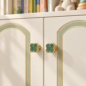 Op de afbeelding: Twee klavervormige kastknoppen met een gladde, donkergroene marmeren inleg, omringd door een gepolijste goudkleurige metalen rand. Deze decoratieve meubelbeslagstukken zijn gemonteerd op een crèmekleurige kastdeur met een gebogen groene rand.