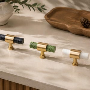 May include: Three cabinet knobs with cylindrical stone handles in black, green, and white. Each knob has a gold-coloured metal base and a cylindrical handle. The knobs are arranged on a light-coloured surface, showcasing their design and materials.