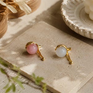 May include: Two gold-colored earrings, each featuring a round gemstone. One earring has a pink gemstone, and the other has a white gemstone. The earrings are displayed on a light-colored surface.