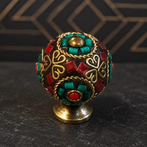 May include: A spherical cabinet knob featuring a dark brown base decorated with inlaid turquoise and red rectangular tiles, gold-toned metal rope detailing, and heart-shaped wire motifs. The knob is mounted on a circular brass-toned metal base.