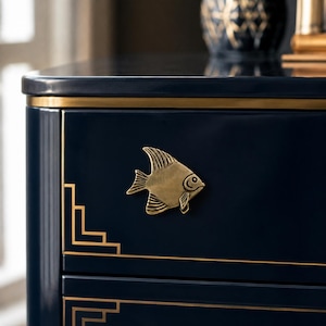 May include: A dark navy blue cabinet drawer featuring a metallic gold fish-shaped handle. The drawer front is accented with thin, linear gold trim detailing along the edges and corners, creating a geometric frame around the central fish hardware.