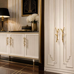 May include: A set of gold-toned metal cabinet handles featuring a slender, branch-like design with delicate leaf accents. These decorative pulls are mounted on white, vertically-ribbed cabinet doors, providing a metallic contrast to the light finish.