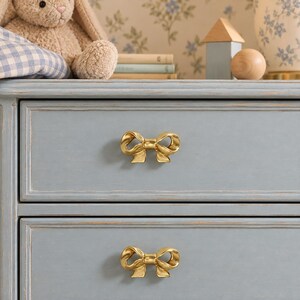 May include: Two gold-toned metal drawer pulls shaped like decorative bows are mounted on the front of a light blue wooden chest of drawers. The drawer pulls feature a polished finish and a detailed ribbon design, providing a functional handle for each drawer.