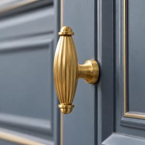 Może przedstawiać: Pionowa gałka meblowa ze szczotkowanego mosiądzu o żłobionym, wrzecionowatym kształcie. Okucie posiada zaokrąglone zakończenia u góry i u dołu, przymocowane do okrągłej podstawy zamontowanej na ciemnoniebieskich drzwiczkach szafki ze złotym wykończeniem.