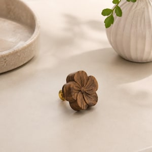 May include: A wooden flower-shaped cabinet knob with a gold-colored base. The knob is carved with detailed petals and is a warm brown color. The knob is likely for furniture or decorative use.