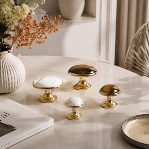Puede incluir: Una colección de tiradores decorativos para armarios con bases doradas y diferentes tapas. Dos tiradores tienen tapas de mármol blanco y dos tienen tapas de piedra ojo de tigre marrón y dorado. También hay un pequeño tirador de mármol. Los tiradores están colocados sobre una mesa de mármol.