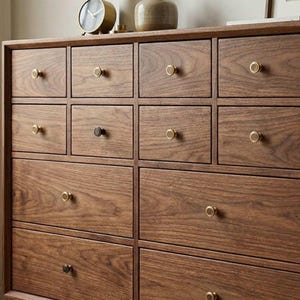 May include: A wooden chest of drawers with numerous square drawers, each fitted with a circular, brass-toned knob. The wood displays a rich grain pattern and a warm brown hue. The design is rectangular and minimalist.