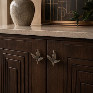 May include: A dark brown wooden cabinet featuring two cabinet doors with recessed paneling and two brass-toned metal handles shaped like stylized leaves. The cabinet is topped with a light beige stone surface.