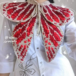 Puede incluir: Un cuello de mariposa hecho a mano con ganchillo en tonos rojos y beige. El cuello presenta un diseño de alas detallado con cierre de lazo. El cuello se lleva sobre una camisa blanca abotonada. El texto "@ellis.crochet" es visible.