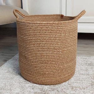 Op de afbeelding: Een cilindrische geweven mand in een natuurlijke bruine kleur. De mand heeft twee kleine handvatten en is gemaakt van een dik, getextureerd materiaal. De mand staat op een lichtgekleurd tapijt.