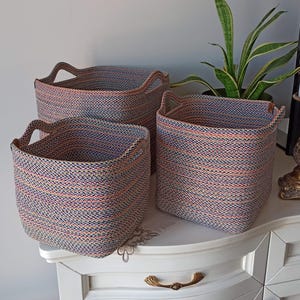 May include: Three woven storage baskets in different sizes. The baskets are made of a blue, orange, and white rope-like material. Each basket has two handles. The baskets are on a white surface.