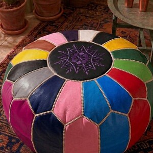 Puede incluir: Un puf marroquí redondo y multicolor con un diseño geométrico. El puf presenta un centro negro con un diseño bordado en morado. Los paneles circundantes son de varios colores, incluyendo rosa, azul, verde y amarillo. Las costuras están acentuadas con un ribete de color claro.