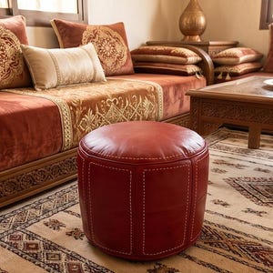 May include: A cylindrical red leather ottoman featuring visible white stitched seams, positioned in front of a wooden sofa with rust-coloured velvet cushions and intricate gold embroidery, set upon a patterned beige and brown area rug.