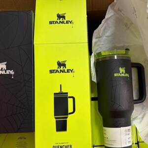 May include: A black Stanley Quencher travel mug with a handle and a green lid, beside yellow and black boxes featuring the Stanley logo and a black box with a spiderweb design.