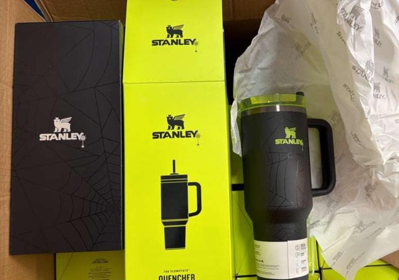 May include: A black Stanley Quencher travel mug with a handle and a green lid, beside yellow and black boxes featuring the Stanley logo and a black box with a spiderweb design.
