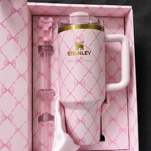 May include: A pink Stanley tumbler with a handle, straw, and lid, presented in a matching gift box. The tumbler and box feature a bow pattern and the text "STANLEY LOVES FANCY". The tumbler has a gold band at the top.