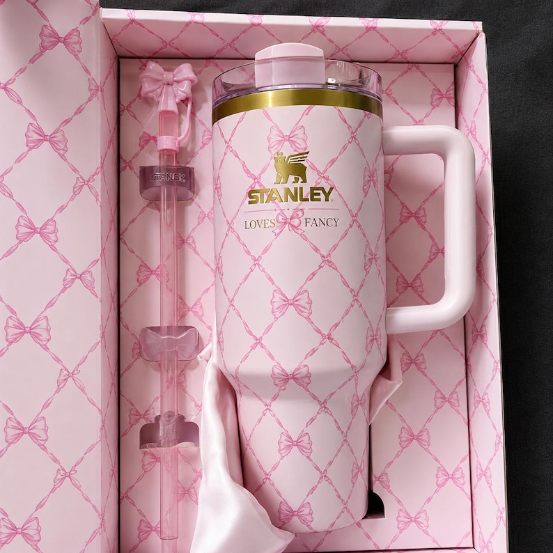 May include: A pink Stanley tumbler with a handle, straw, and lid, presented in a matching gift box. The tumbler and box feature a bow pattern and the text "STANLEY LOVES FANCY". The tumbler has a gold band at the top.