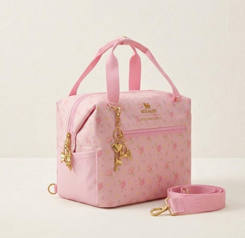 May include: A pink floral Stanley bag with a top handle and a detachable shoulder strap. The bag features gold-colored hardware, including a zipper, charms, and a clasp. The text "STANLEY LOVEISRADIANCE" is printed on the front.