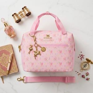 May include: A pink floral print handbag with a matching shoulder strap, featuring a top handle and a gold-tone zipper. The bag is adorned with gold charms and is surrounded by a perfume bottle, a journal, and a pair of binoculars.