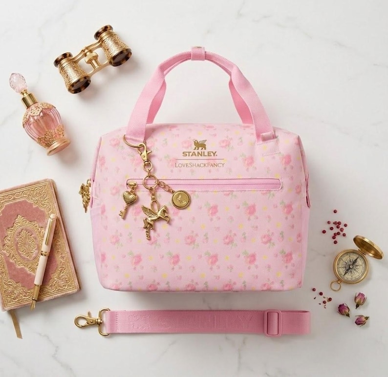 May include: A pink floral print handbag with a matching shoulder strap, featuring a top handle and a gold-tone zipper. The bag is adorned with gold charms and is surrounded by a perfume bottle, a journal, and a pair of binoculars.
