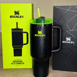 May include: Black Stanley Quencher H2.0 tumbler with a green lid and straw. The tumbler has a handle and is next to a bright green box and a black box. The tumbler is 1.18L.
