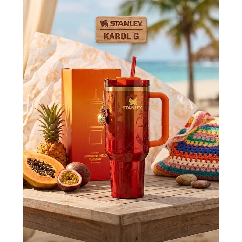 May include: A red Stanley Quencher H2.0 tumbler with a gold logo, orange handle, and matching straw. The tumbler features a floral pattern and a small pineapple charm. It is displayed next to its orange box, a pineapple, and a halved papaya.