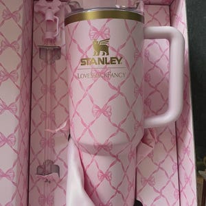 May include: A pink Stanley Loveshackfancy tumbler with a handle, straw, and lid, presented in a matching box. The tumbler and box feature a repeating bow pattern. The Stanley logo is visible on the tumbler.