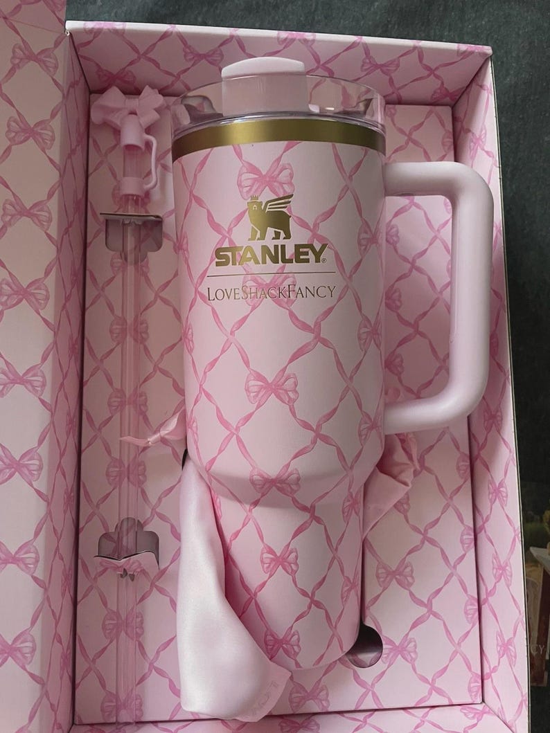 May include: A pink Stanley Loveshackfancy tumbler with a handle, straw, and lid, presented in a matching box. The tumbler and box feature a repeating bow pattern. The Stanley logo is visible on the tumbler.