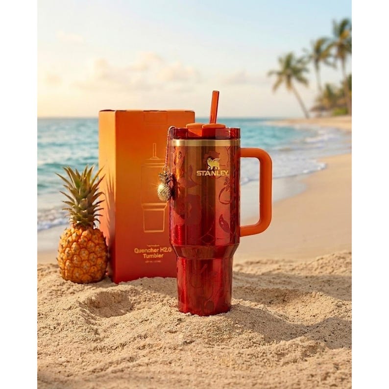 May include: A red Stanley Quencher H2.0 tumbler with a matching orange handle, straw, and lid. The tumbler features a subtle floral pattern, a gold Stanley logo, and a small pineapple charm attached to the lid. Beside it stands its orange box.