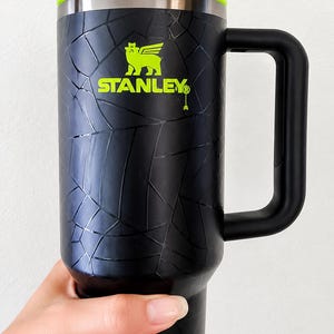 May include: Black Stanley travel mug with a neon green lid and straw. The mug features a black handle and the Stanley logo in green. The exterior has a textured pattern.