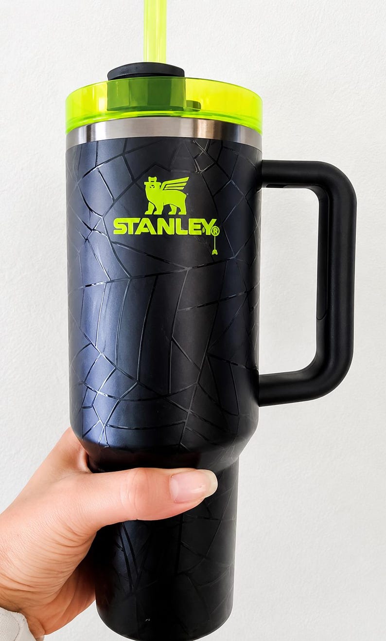 May include: Black Stanley travel mug with a neon green lid and straw. The mug features a black handle and the Stanley logo in green. The exterior has a textured pattern.