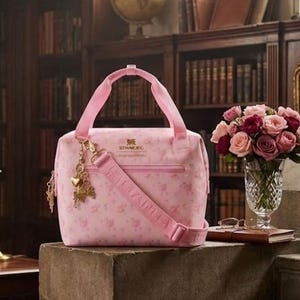 May include: A pink floral-patterned handbag with a matching shoulder strap and handles. The bag features a gold charm and a zipper closure. A bouquet of pink and red roses sits in a crystal vase nearby. The bag has the text "STHMEG".