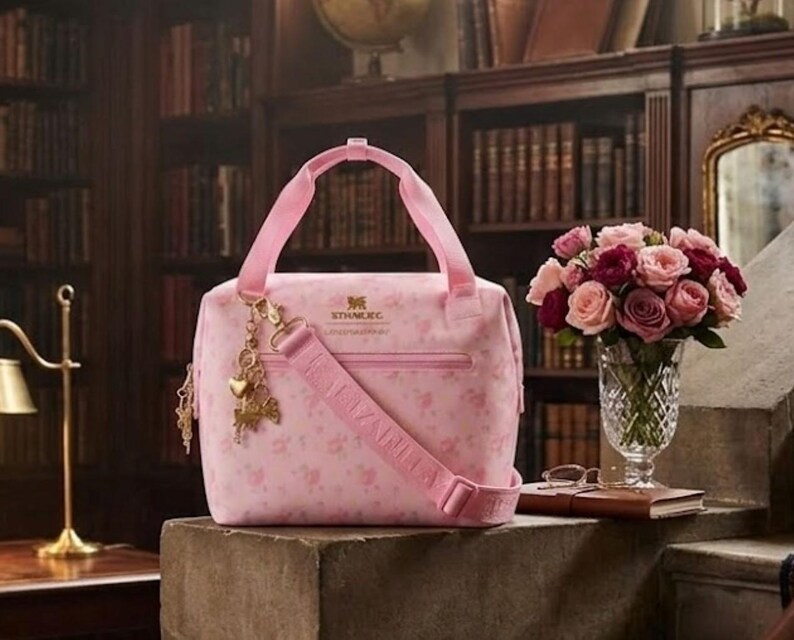 May include: A pink floral-patterned handbag with a matching shoulder strap and handles. The bag features a gold charm and a zipper closure. A bouquet of pink and red roses sits in a crystal vase nearby. The bag has the text "STHMEG".