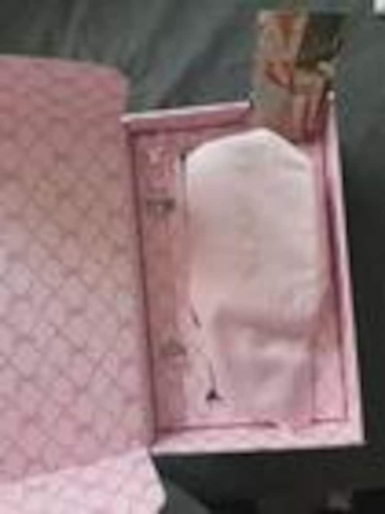 May include: A pink gift box with a diamond pattern on the inside and a white object wrapped in pink tissue paper. The box is open, revealing the contents. The background is a dark gray surface.