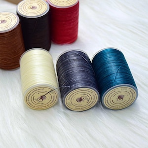 May include: A collection of spools of sewing thread in various colors, including brown, black, red, cream, navy, and teal. Each spool has a paper label with text. The threads are arranged on a white, fluffy surface.
