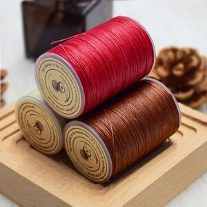 May include: Three spools of sewing thread in red, brown, and cream colors. The spools are stacked on a wooden tray. Each spool has a label with text. The thread is tightly wound, and the colors are vibrant.