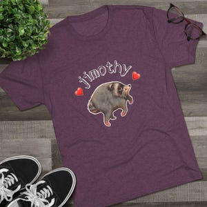 May include: A heathered purple short-sleeve t-shirt featuring a central graphic of a round, grey raccoon-like creature. Above the creature, the text Jimothy is printed in white, flanked by two small red hearts on either side of the illustration.