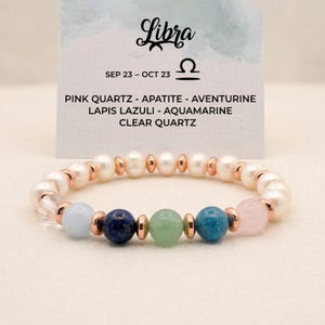 May include: A Libra zodiac bracelet featuring a mix of gemstones and pearls. The bracelet includes pink quartz, apatite, aventurine, lapis lazuli, aquamarine, and clear quartz beads, with rose gold accents. The text on the card reads "Libra" and "SEP 23 - OCT 23".