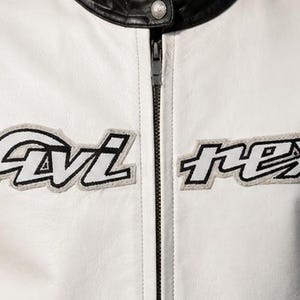 May include: Close-up of a white leather jacket with black accents. The jacket features the embroidered text "Avi rex" in black and white. A black zipper runs down the center, and the collar is black.